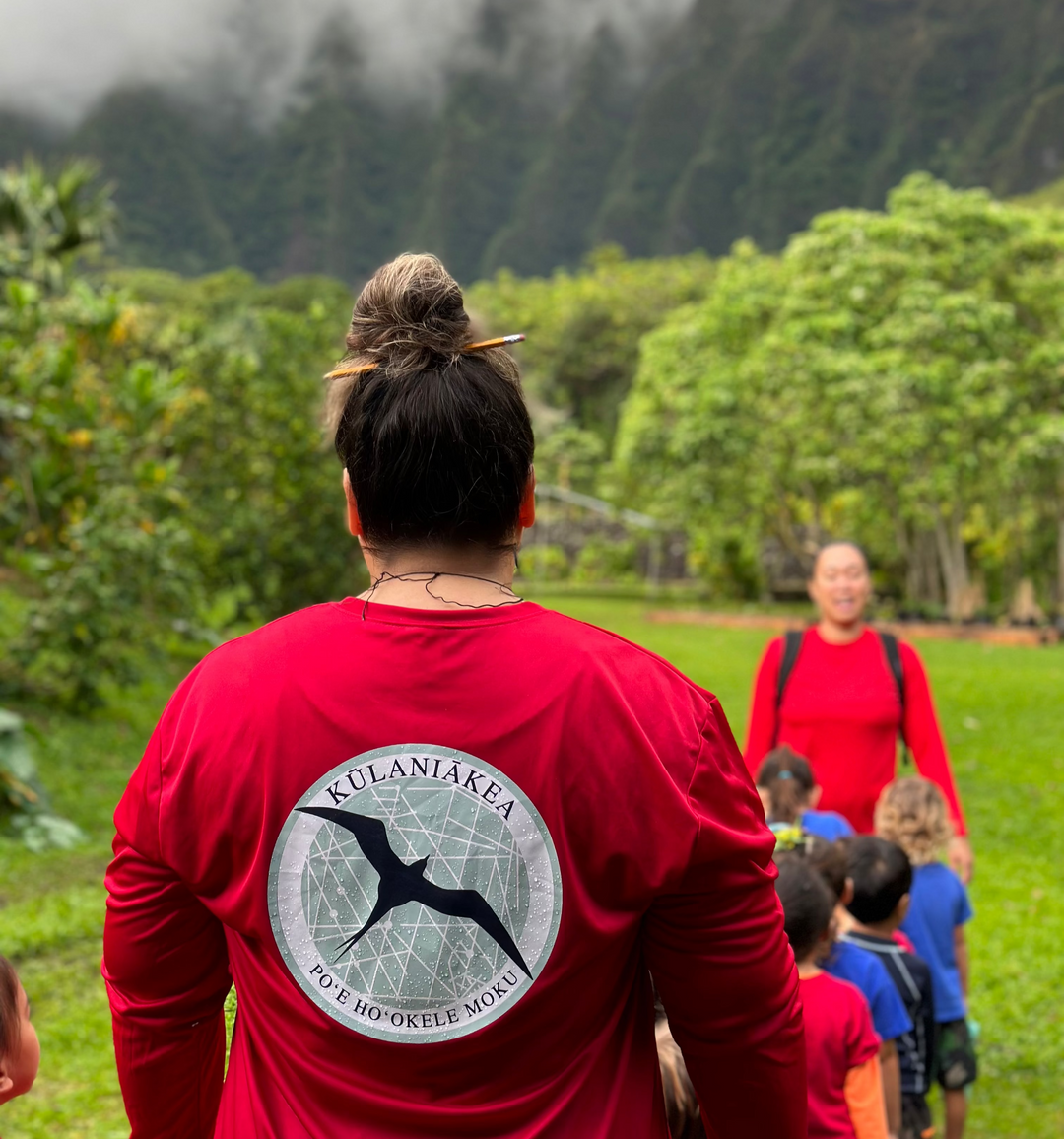 Person wearing a red shirt with a kulaniakea logo and text, standing in a natural setting with trees and mountains.