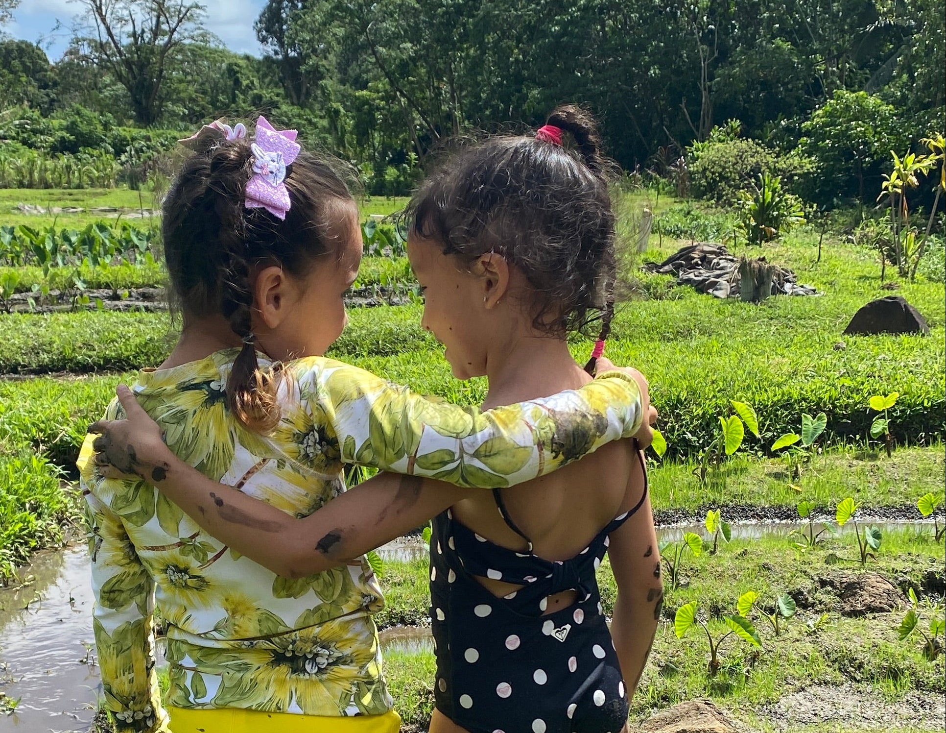 Two young girls standing in muddy grass outdoors, wearing swimwear and looking at each other. One wears a yellow patterned long-sleeve top and yellow shorts, and the other wears a black polka-dot swimsuit.