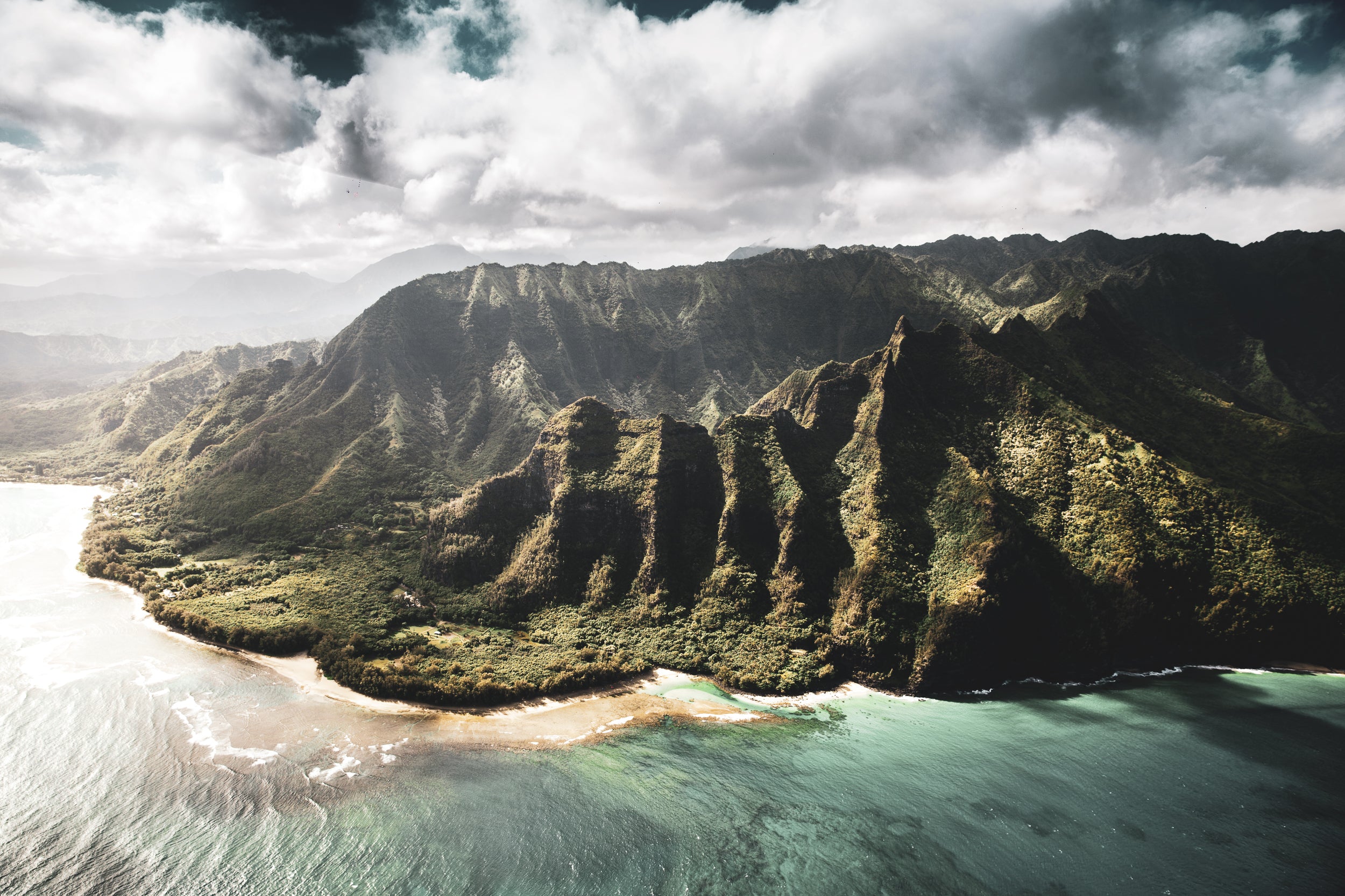 Aerial view of a dramatic coastal landscape with steep green mountains, turquoise water, a sandy shoreline, and a cloudy sky.